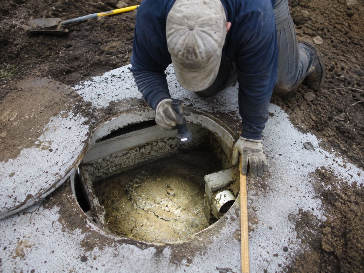 Technician kneeling at an open septic tank, checking the outlet with a flashlight and tape measure