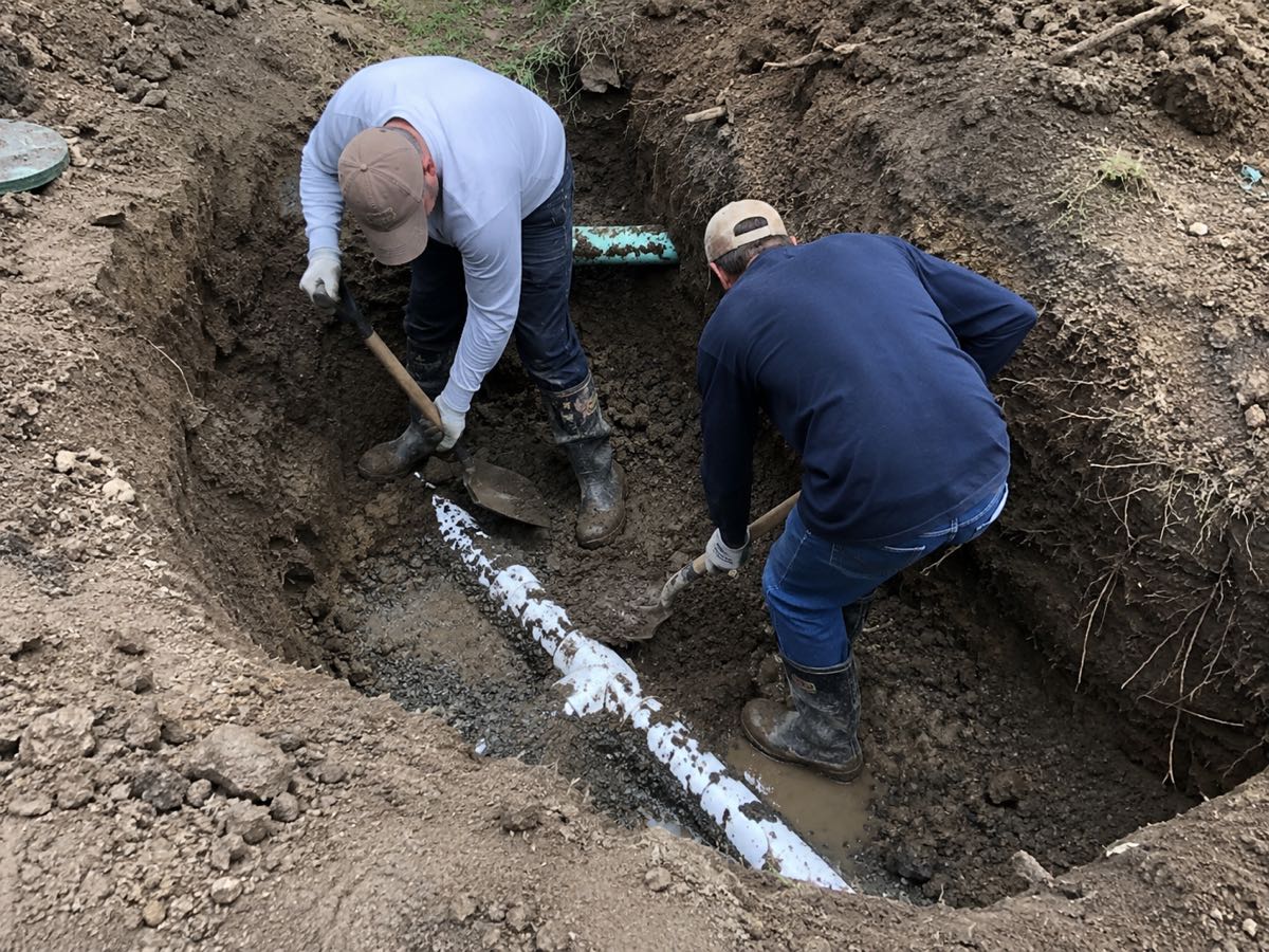Two workers setting a repaired septic line into a gravel trench by hand